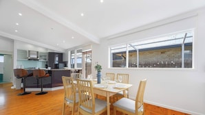 A dining area with a wooden table and chairs, a kitchen in the background, and a large window with a view. - 4BR Marsfield Stay  Walk to Macquarie Uni Metro (Marsfield)