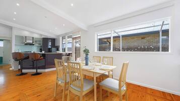 A dining area with a wooden table and chairs, a kitchen in the background, and a large window with a view.