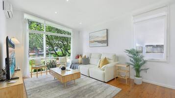 A modern living room with a sofa, coffee table, and a large window offering a view of greenery outside.
