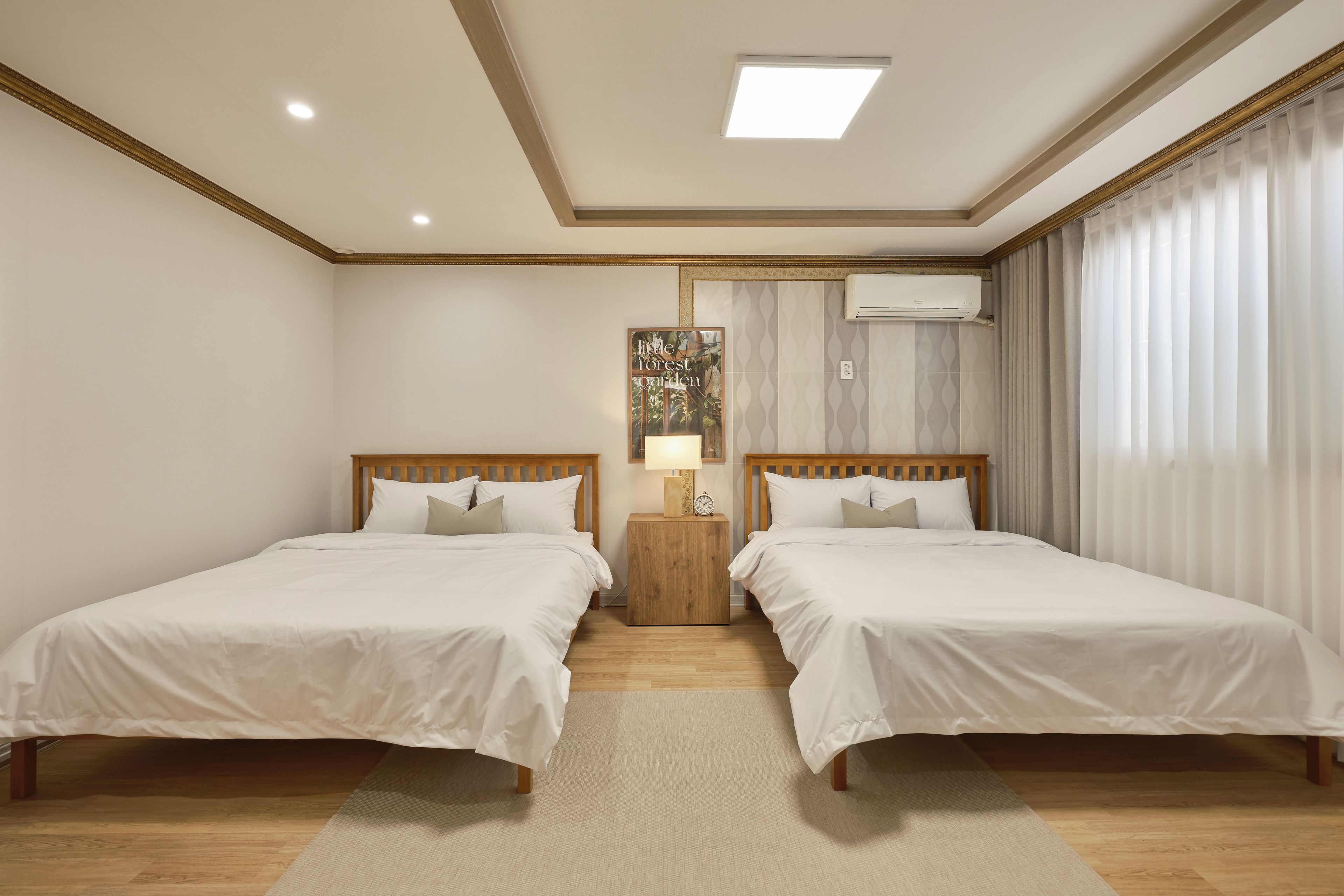 A hotel room with two beds, a nightstand with a lamp, an air conditioning unit, and a window with curtains.