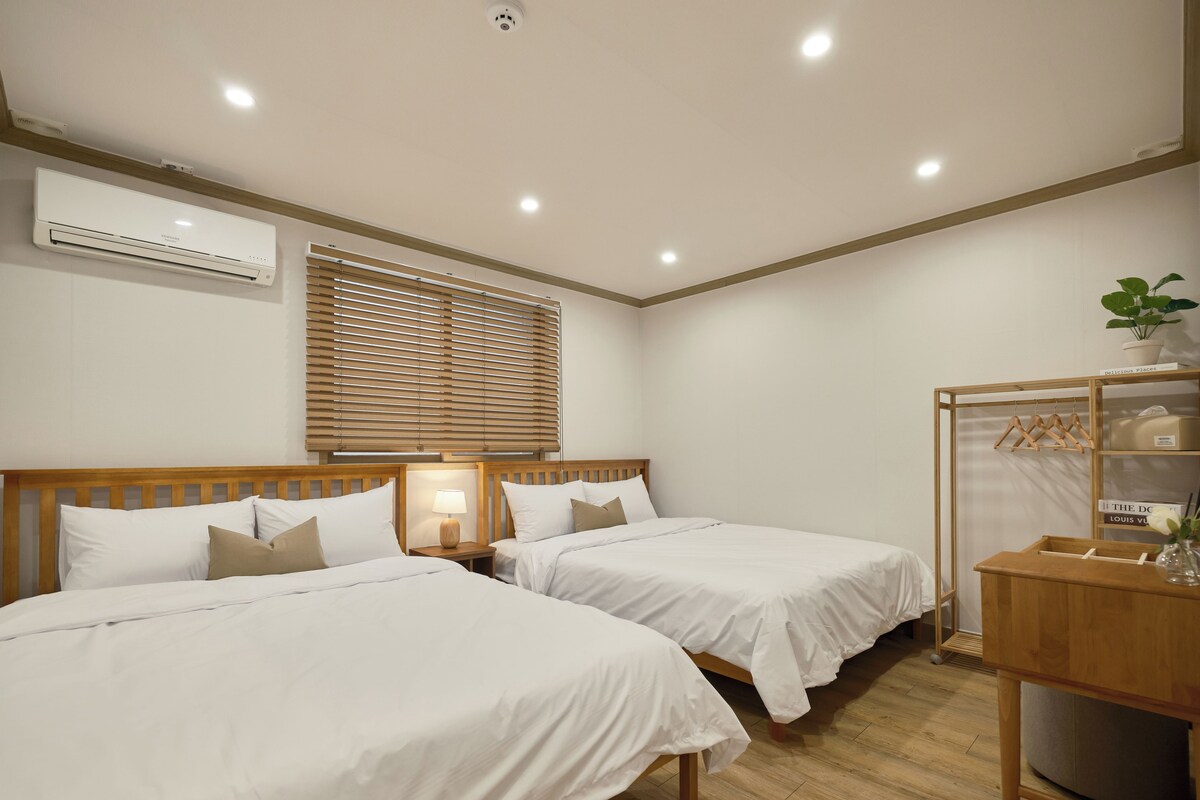 A hotel room with two beds, a bedside table with a lamp, a wooden headboard, a window with blinds, a wardrobe, and a potted plant.