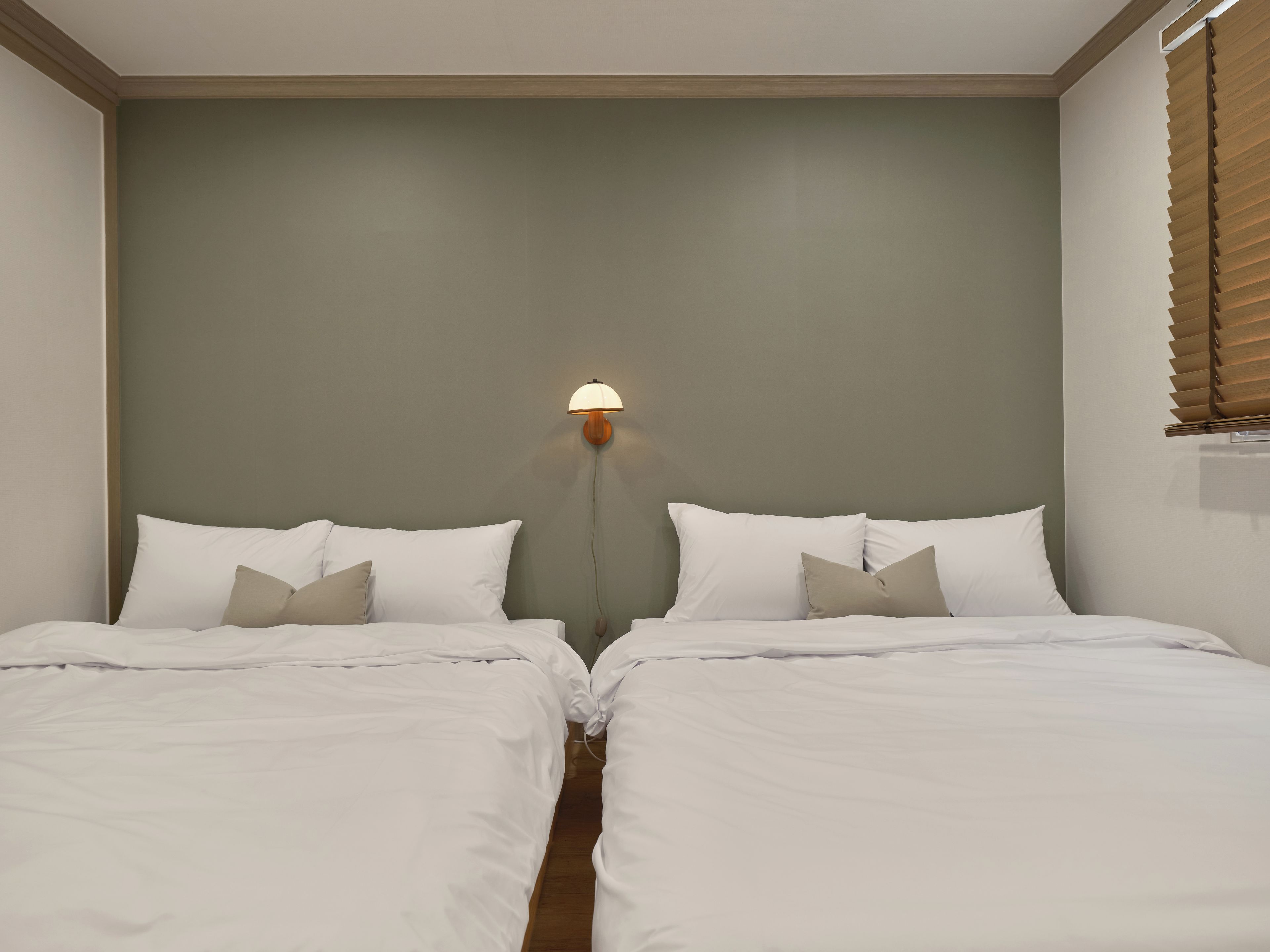 Ein Doppelbett mit weißer Bettwäsche und zwei Kissen, eine Wandleuchte und ein Fenster mit Jalousien.