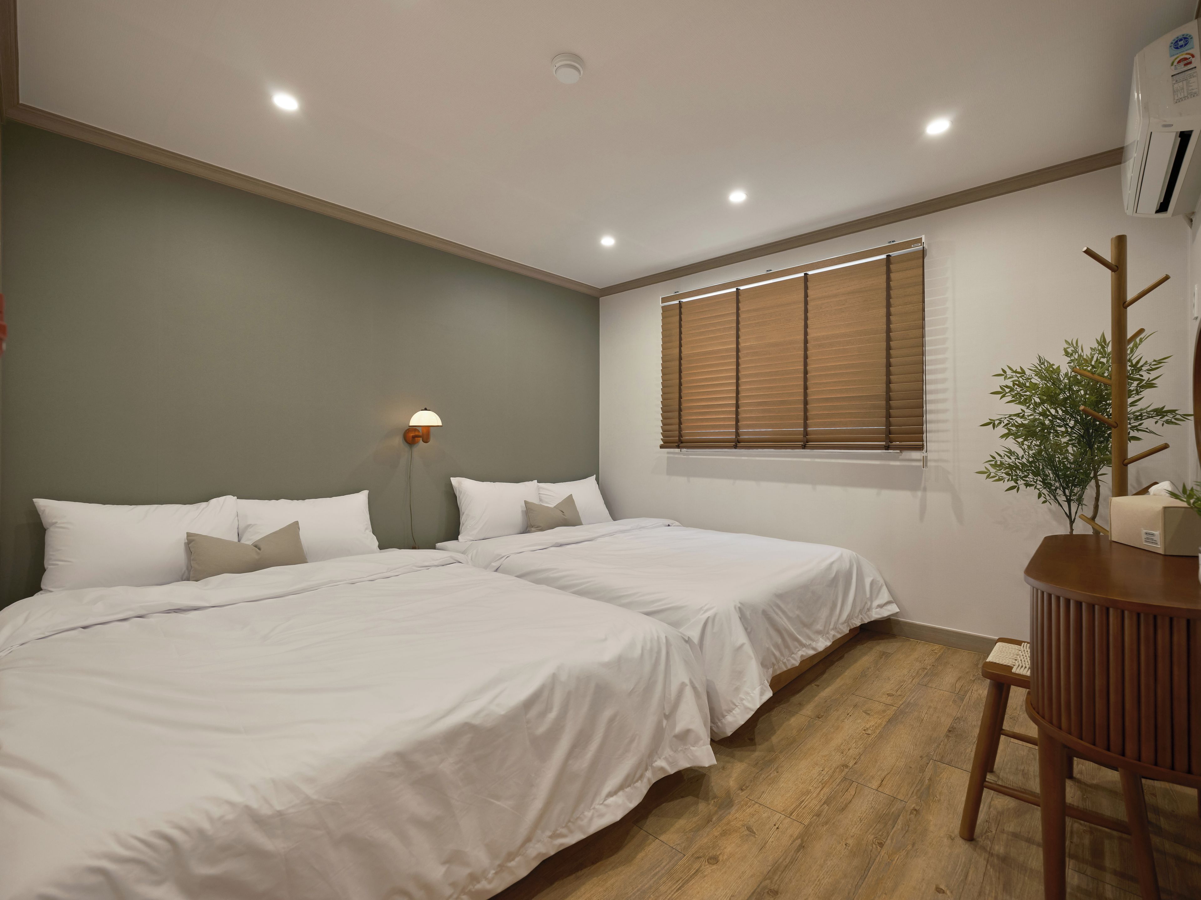 A bedroom with two beds, a bedside table, a lamp, a window with blinds, and an air conditioning unit.