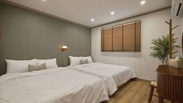 A bedroom with two beds, a bedside table, a lamp, a window with blinds, and an air conditioning unit.