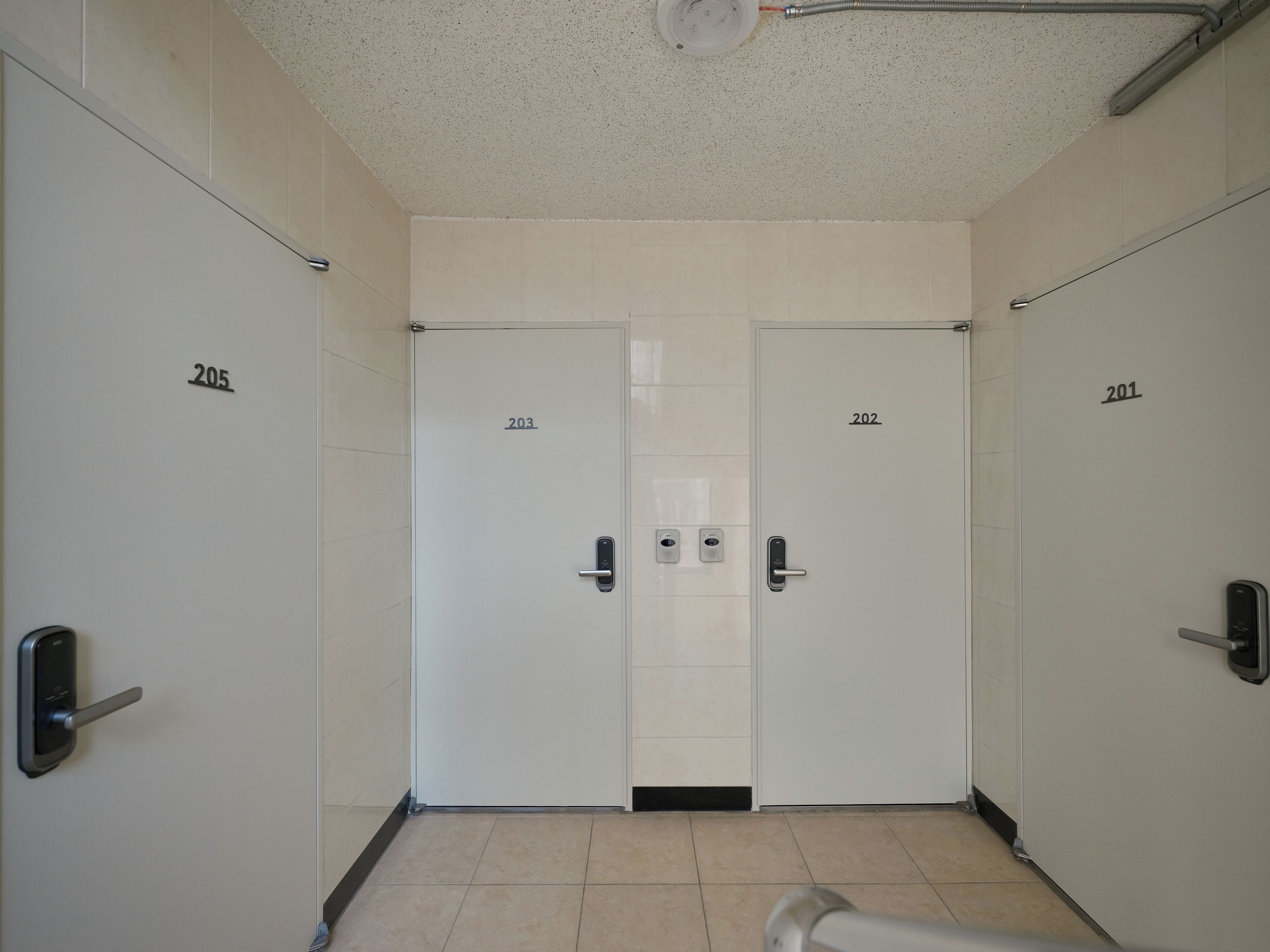 Een gang met twee deuren, waarop respectievelijk de aanduidingen 203 en 202 staan, elk met een moderne deurklink en een klein paneel aan de muur.