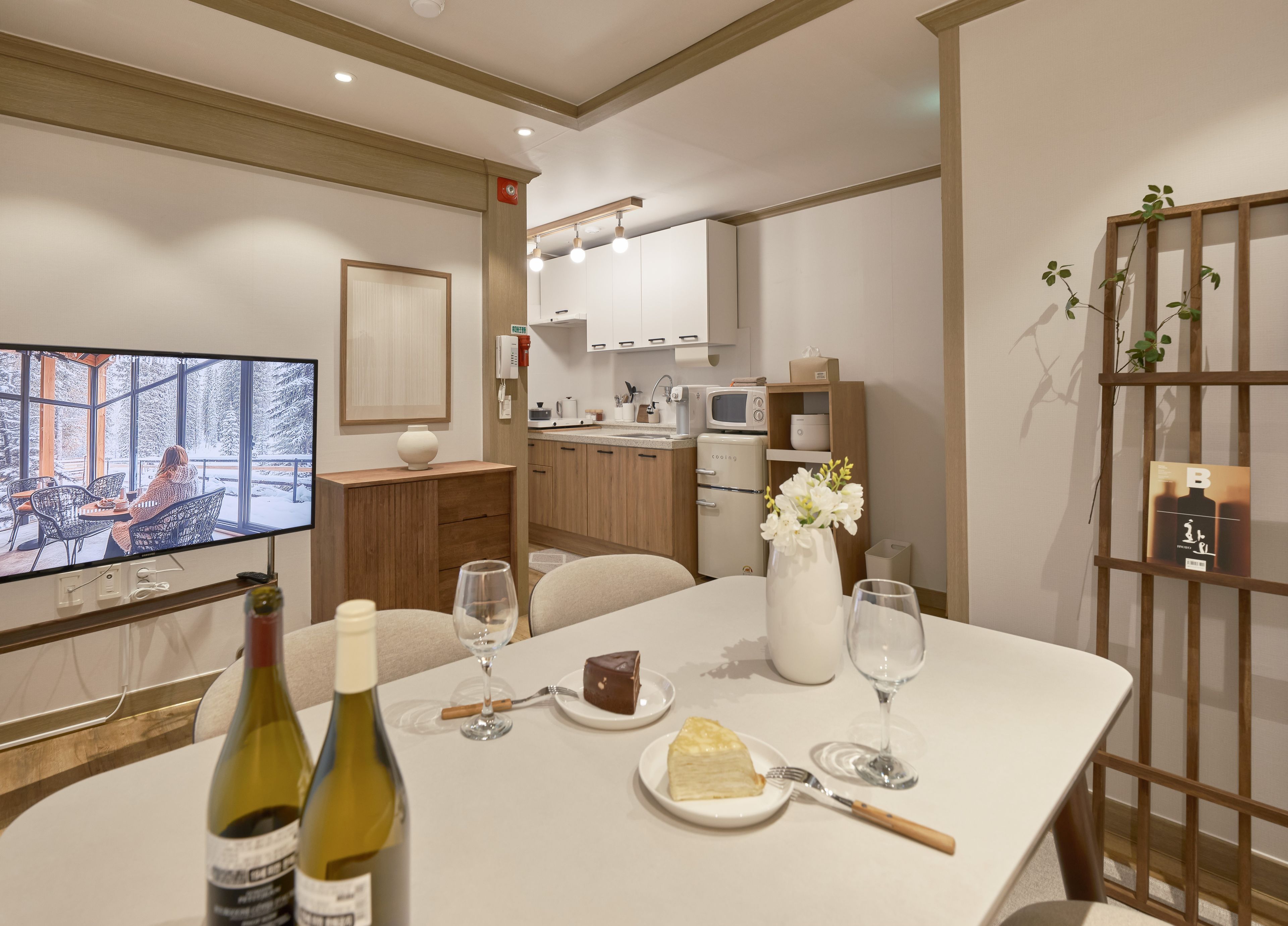 A modern dining area with a television displaying a snowy landscape, a table set with wine glasses, a cake, and a vase with flowers.
