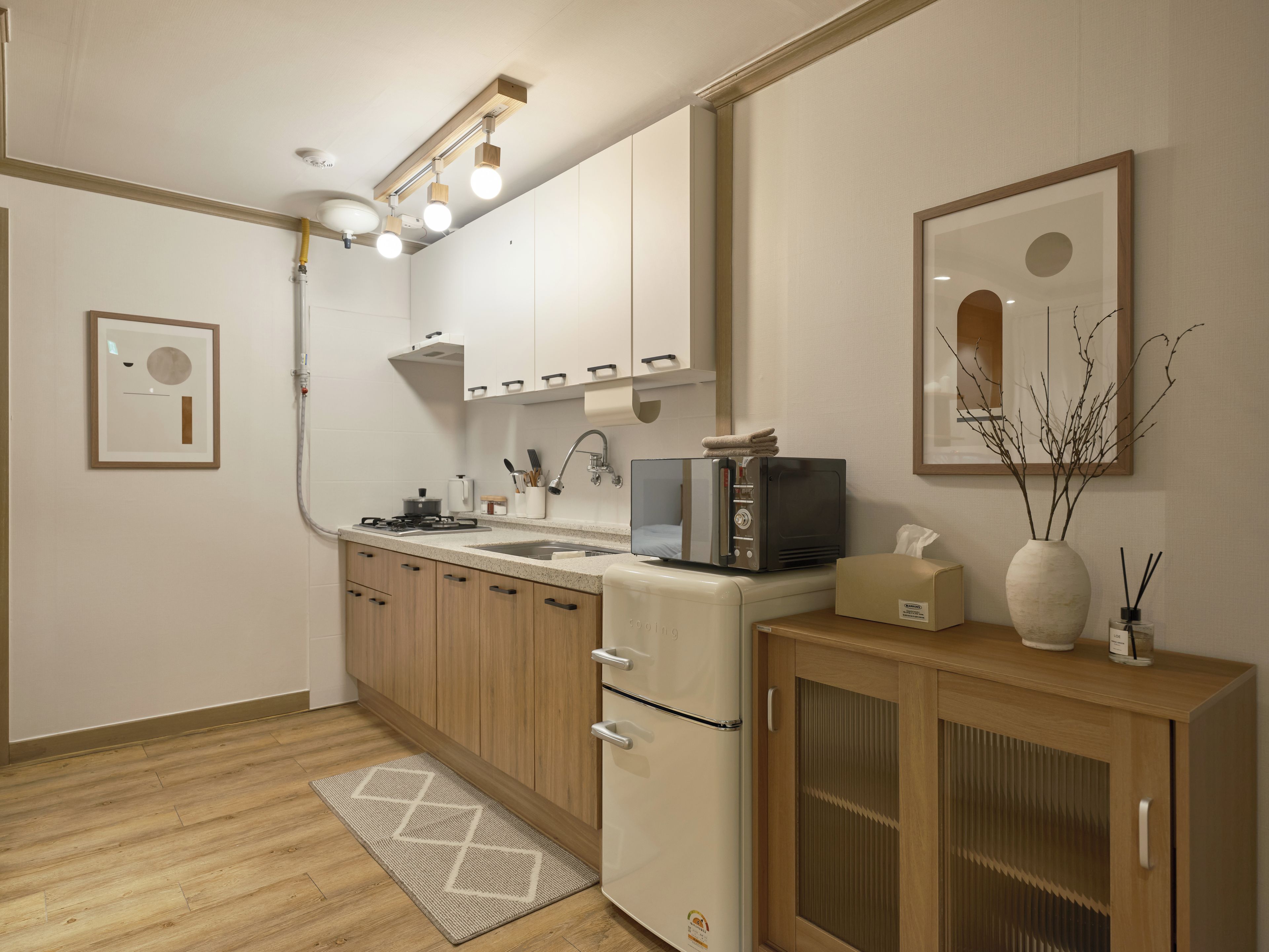 A modern kitchen with a built-in oven, microwave, and sink. There is a wooden cabinet with a glass door and a small refrigerator. A rug is on the floor, and a framed picture hangs on the wall.