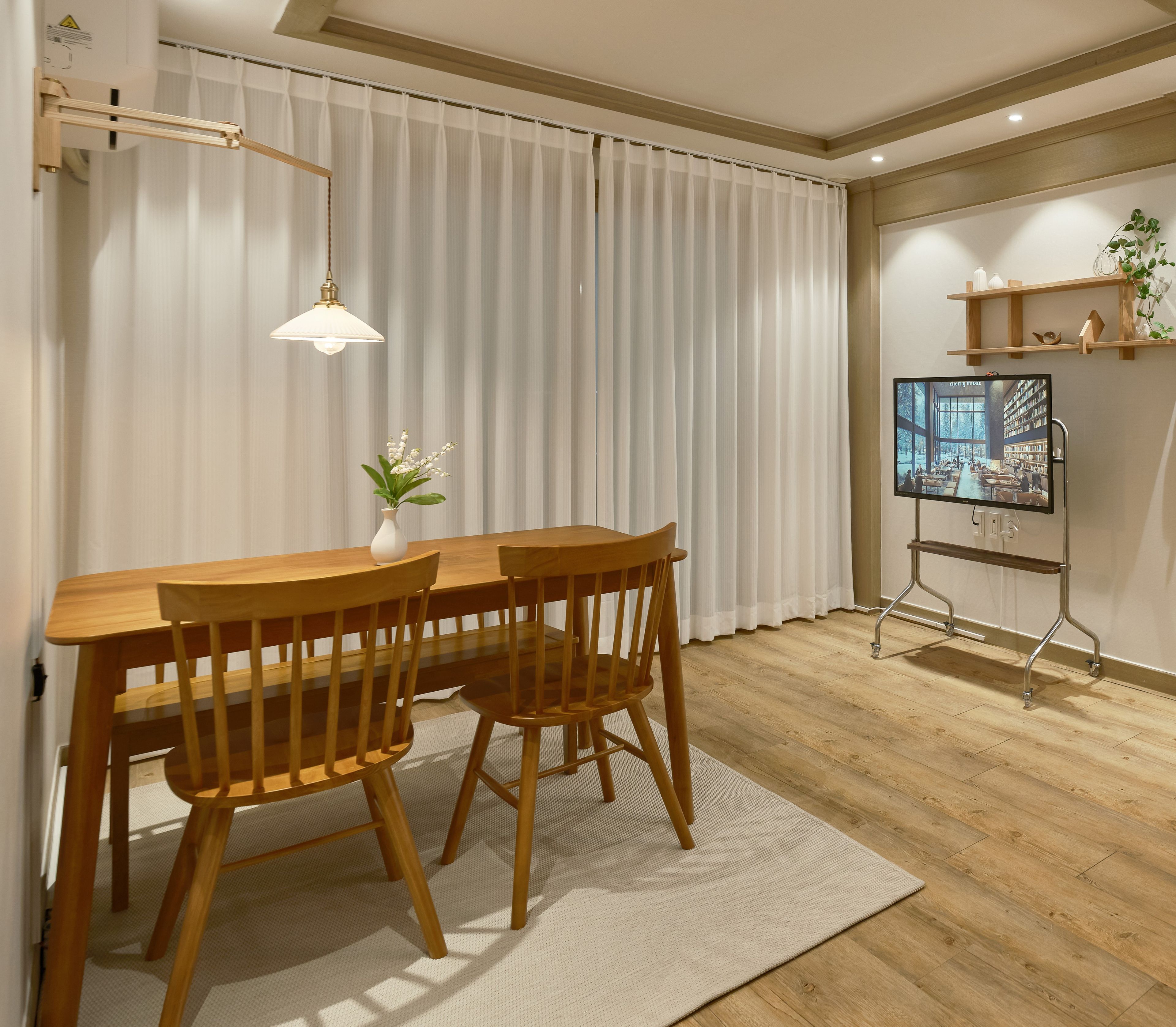 A dining area with a wooden table and chairs, a white vase with flowers, a hanging light fixture, and a television on a stand.