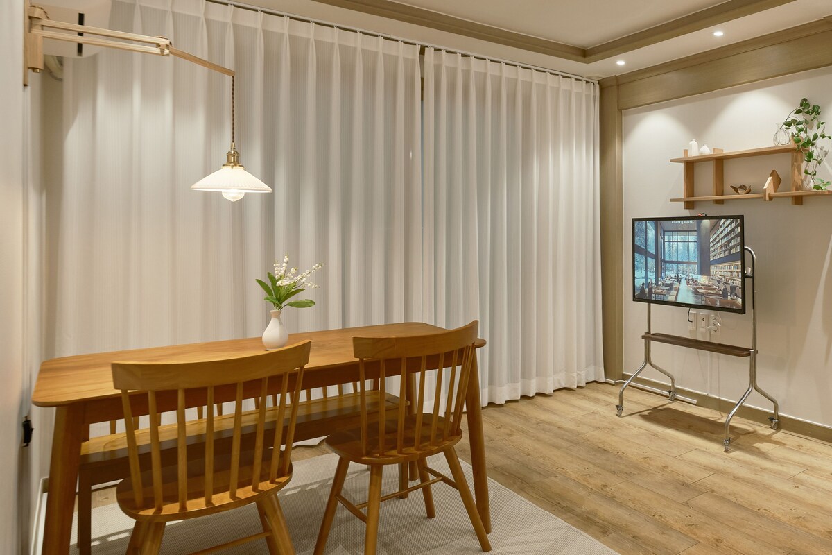A dining area with a wooden table and chairs, a white vase with flowers, a hanging light fixture, and a television on a stand.