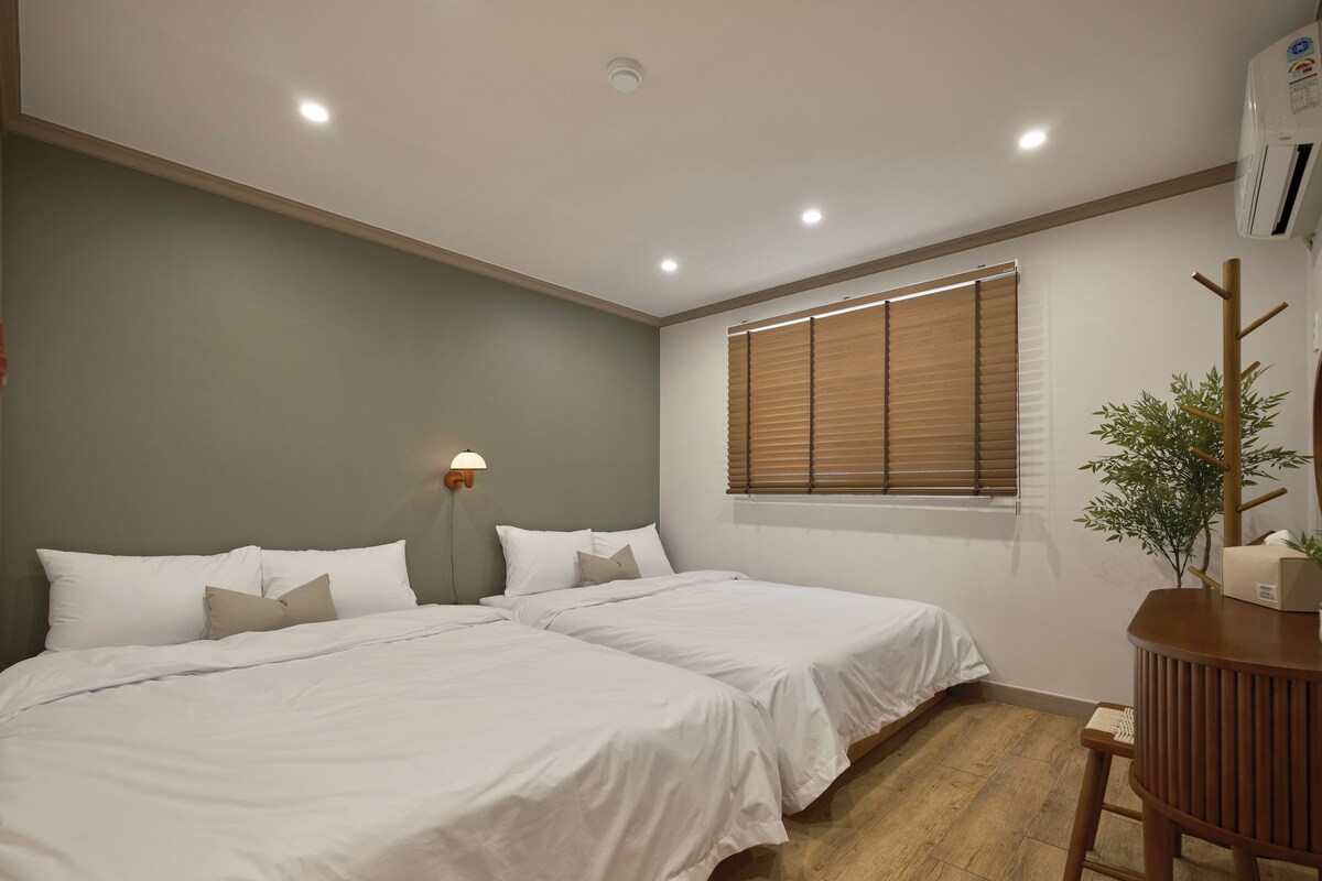 A bedroom with two beds, a bedside table, a lamp, a window with blinds, and an air conditioning unit.