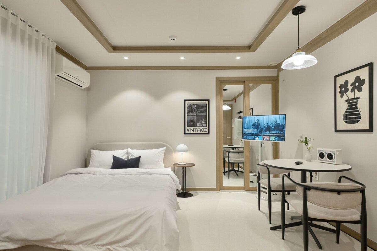 A hotel room with a bed, bedside table, lamp, TV, and a small dining area with chairs and a table.