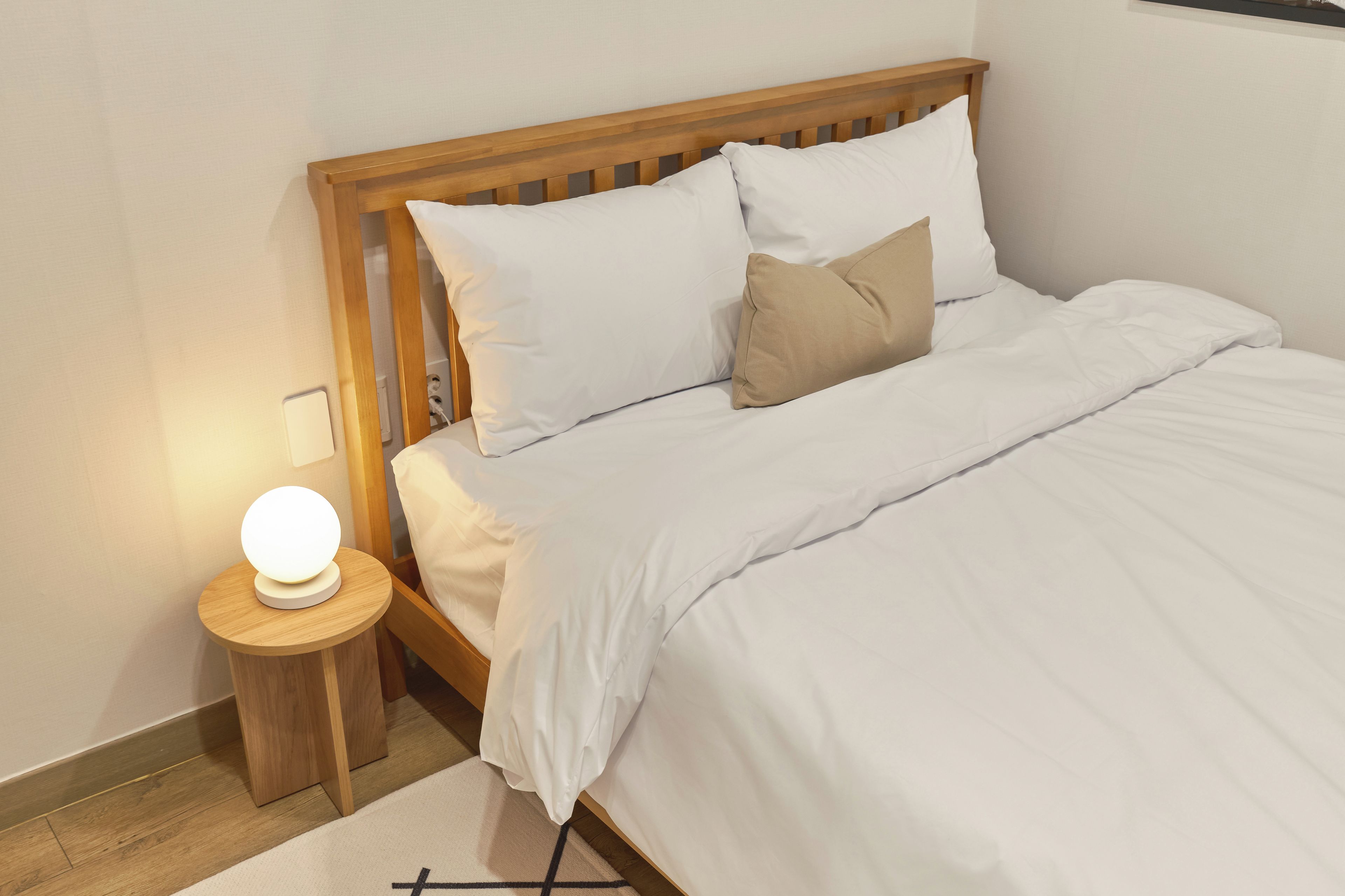 A neatly made bed with white bedding and a single tan pillow, a small bedside table with a round lamp, and a wooden headboard.