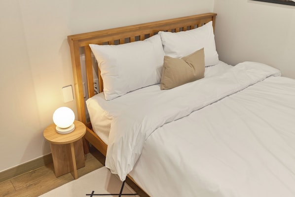 Una cama bien hecha con ropa de cama blanca y una sola almohada color arena, una mesita de noche pequeña con una lámpara redonda y un cabecero de madera.