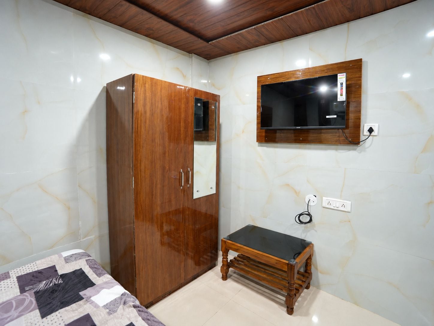 Quarto com guarda-roupa de madeira, um pequeno banco de madeira, televisão fixada na parede e cama com colcha estampada.