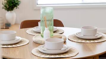 A round dining table with a set of white plates and bowls, a green vase with reed sticks, and a small potted plant in the background.