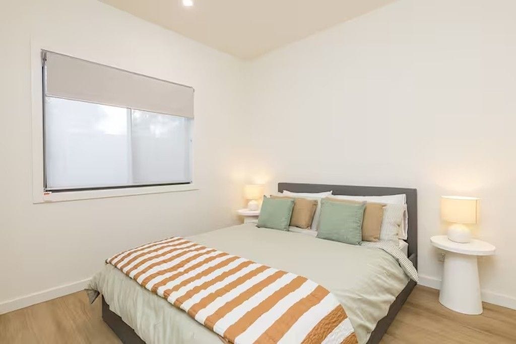 A bedroom with a bed, two bedside tables, a window with blinds, and a wall-mounted lamp.