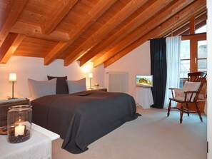 A bedroom with a wooden ceiling, a bed with a dark blanket, a chair, a table with a candle, and a television on a stand. - 1 in Katrin's House (Bad Hindelang)