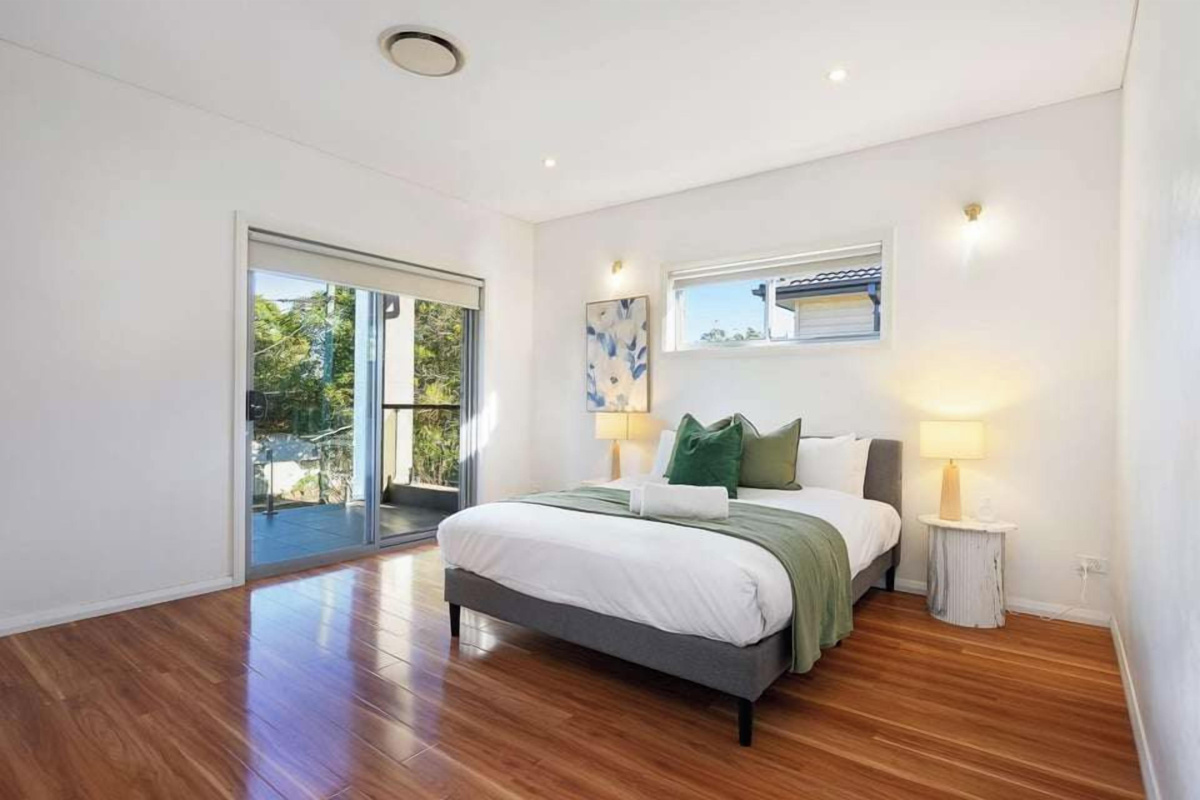 A modern bedroom with a large bed, a sliding glass door leading to a balcony, and a small table with a lamp.
