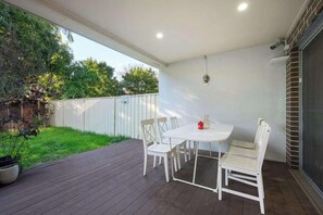 Um pátio externo com mesa e cadeiras brancas, rodeado por um deck de madeira e um cercado branco.