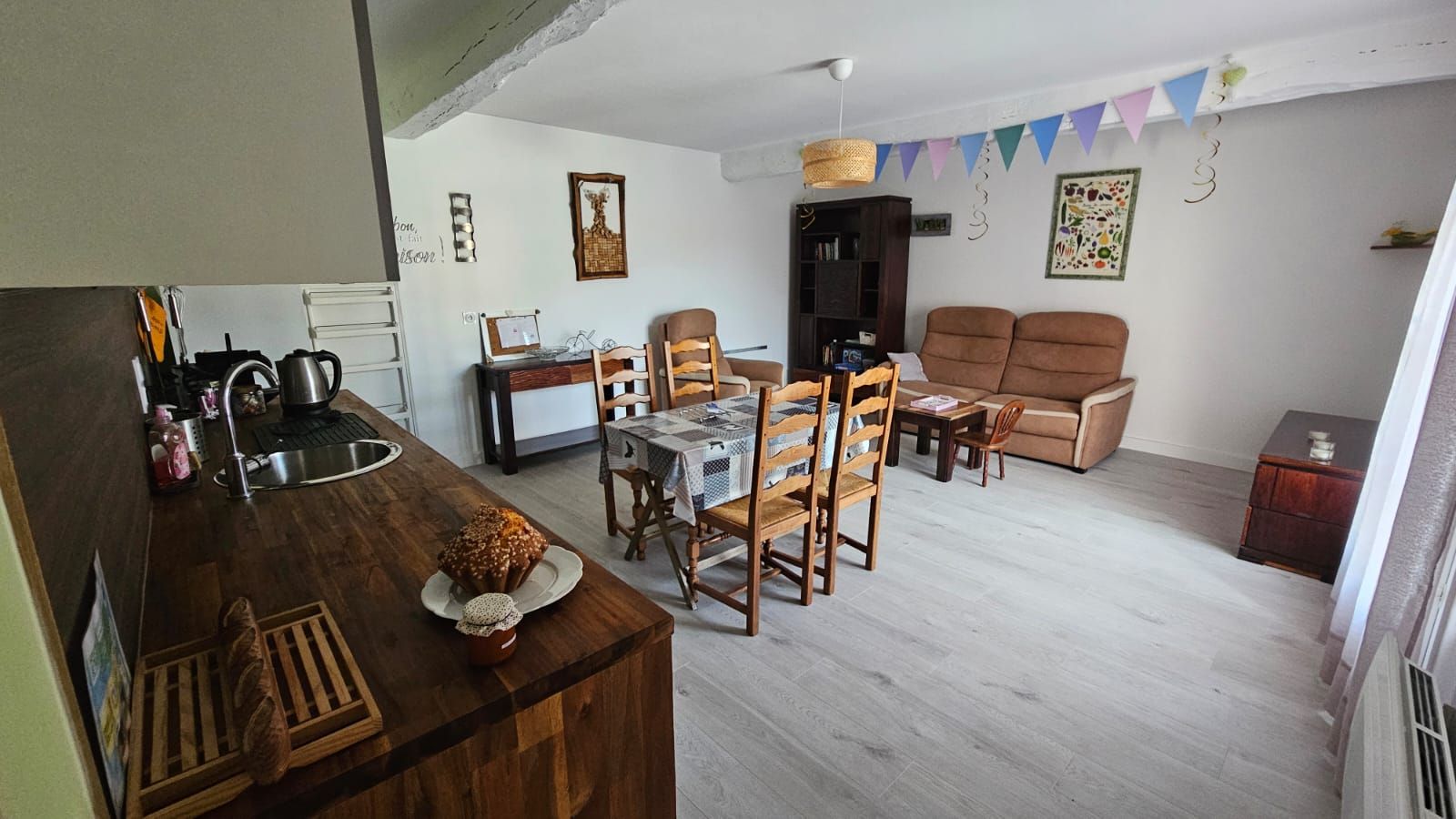 Un salon chaleureux avec une table à manger en bois, deux canapés et un coin cuisine équipé d’un évier et d’une bouilloire.