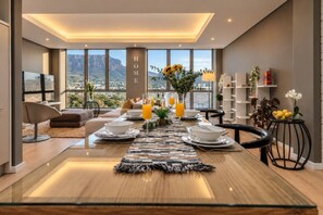 A modern living room with a dining area, large windows, and a view of mountains. - Mandela Rhodes Sky Suite I Mountain & Pool Views (Cape Town)