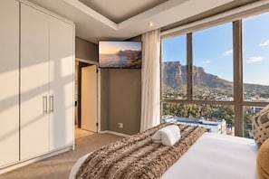 A hotel room with a bed, a TV, a large window with a city view, and a wardrobe. - Mandela Rhodes Sky Suite I Mountain & Pool Views (Cape Town)