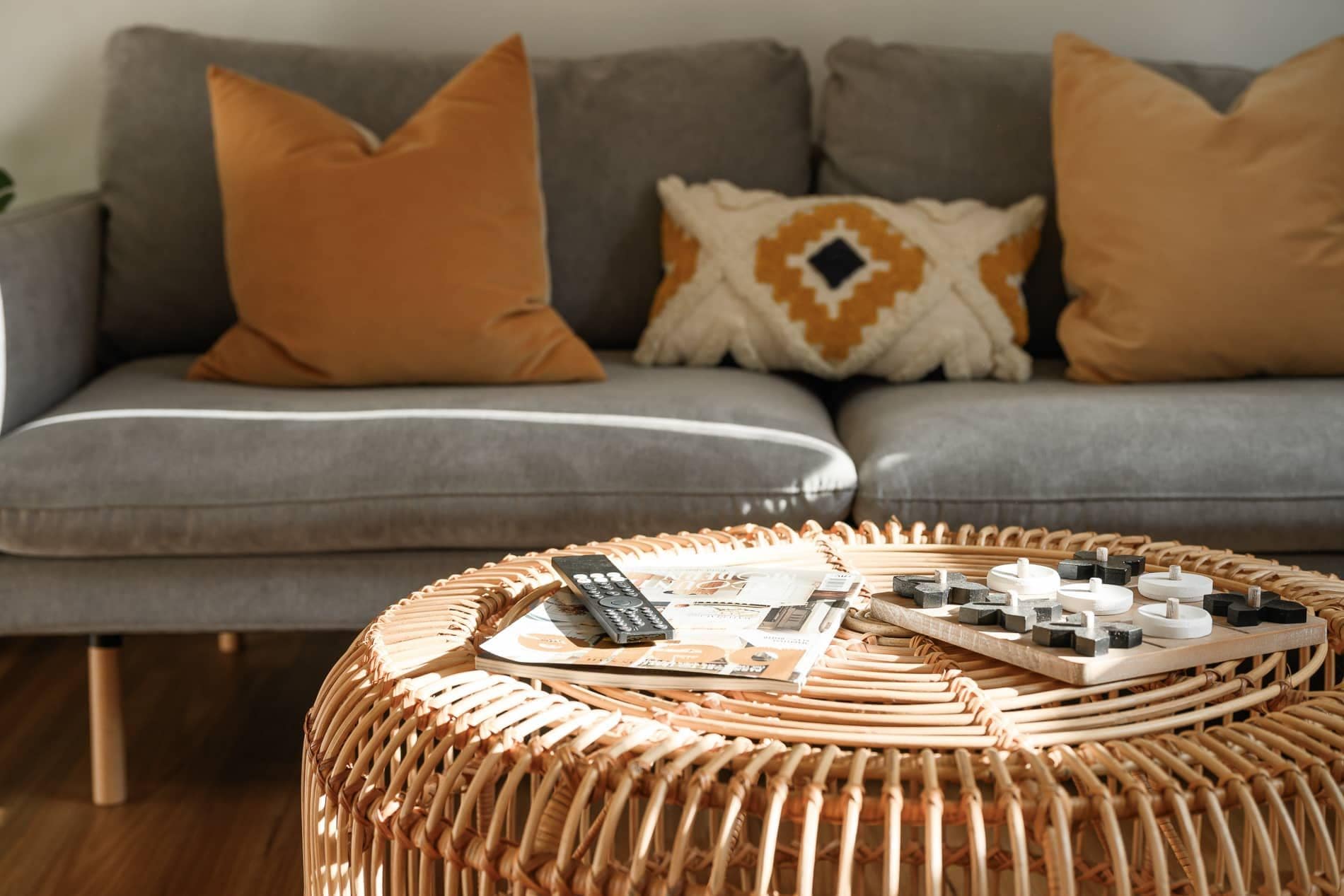 Un canapé gris agrémenté de deux coussins orange et d’un coussin à motifs, une table basse en osier sur laquelle reposent une télécommande, des magazines et de petits objets.