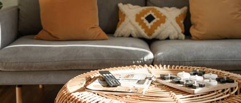 Un canapé gris agrémenté de deux coussins orange et d’un coussin à motifs, une table basse en osier sur laquelle reposent une télécommande, des magazines et de petits objets.