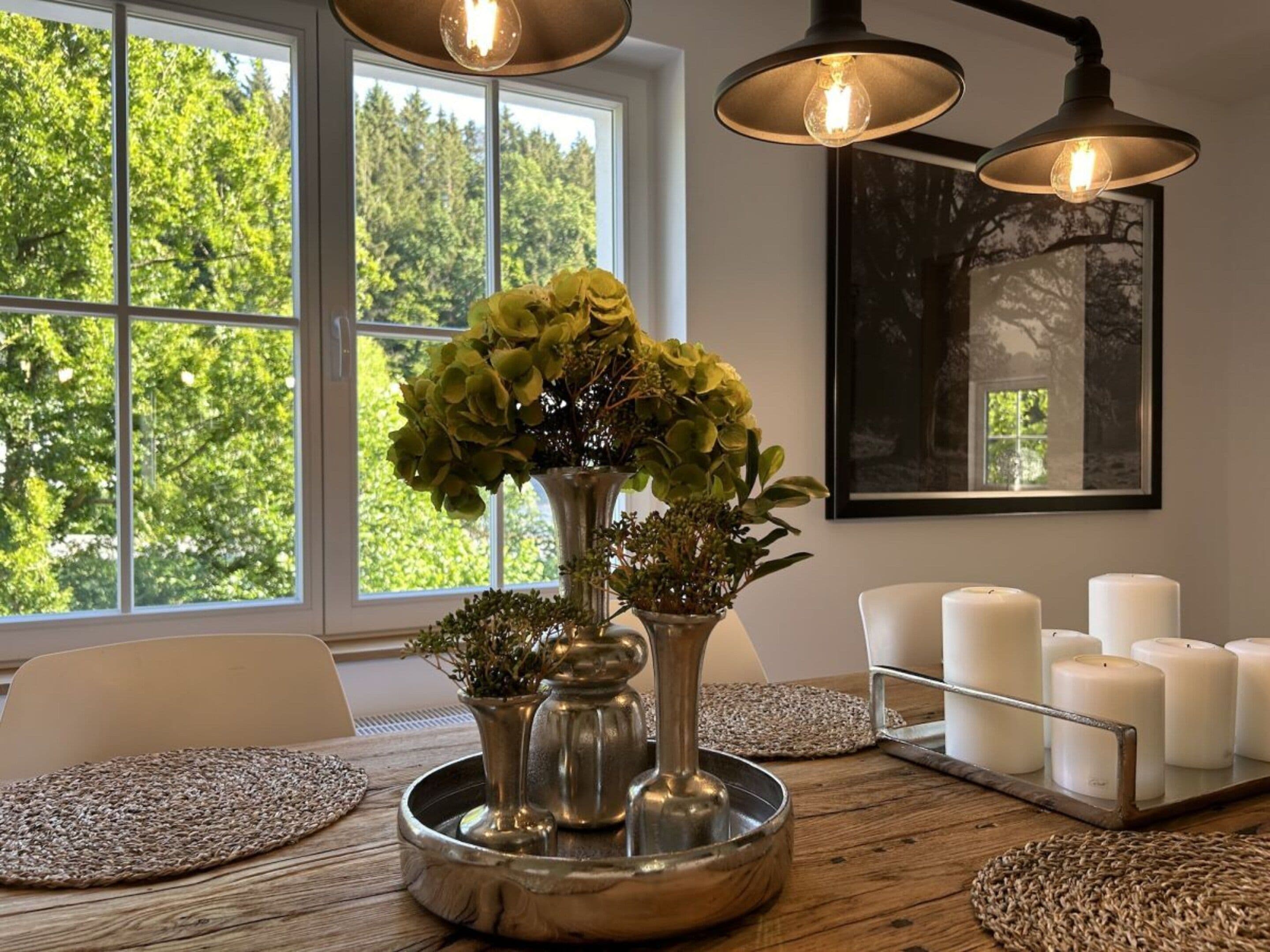 A room with a dining table, a vase with green plants, and a candle holder with white candles.