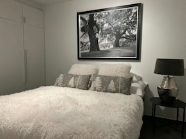 A bedroom with a white fluffy bed, two pillows with tree patterns, a framed black and white photograph on the wall, and a lamp on a side table.