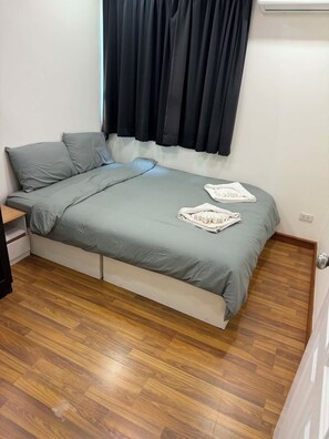 Ein Einzelbett mit grauem Matratze und Kissen, Holzboden und ein Fenster mit dunklen Vorhängen.