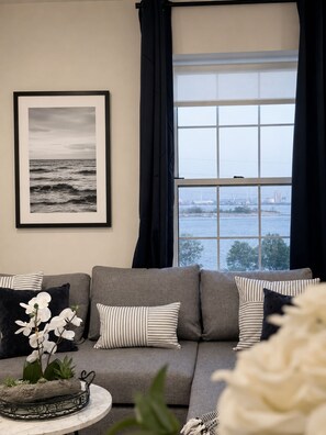 Ein Wohnzimmer mit grauem Sofa, gestreifter Dekokissen, einem Schwarz-Weiß-Foto und Blick auf ein Gewässer durch ein Fenster.
