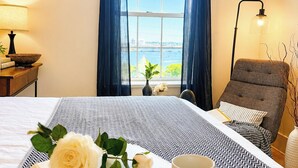 Une chambre d’hôtel avec un lit, un coin lecture comprenant un livre et une tasse, une chaise, une lampe et une vue sur la mer par la fenêtre.