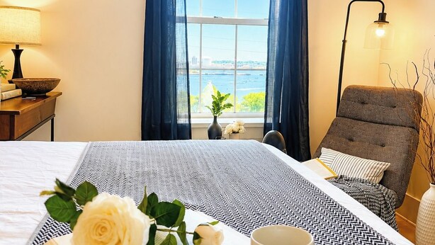 A hotel room with a bed, a reading area with a book and a cup, a chair, a lamp, and a view of the sea through a window. - Water View 1BR near Brown & RISD, Parking + Elevator (Providence)