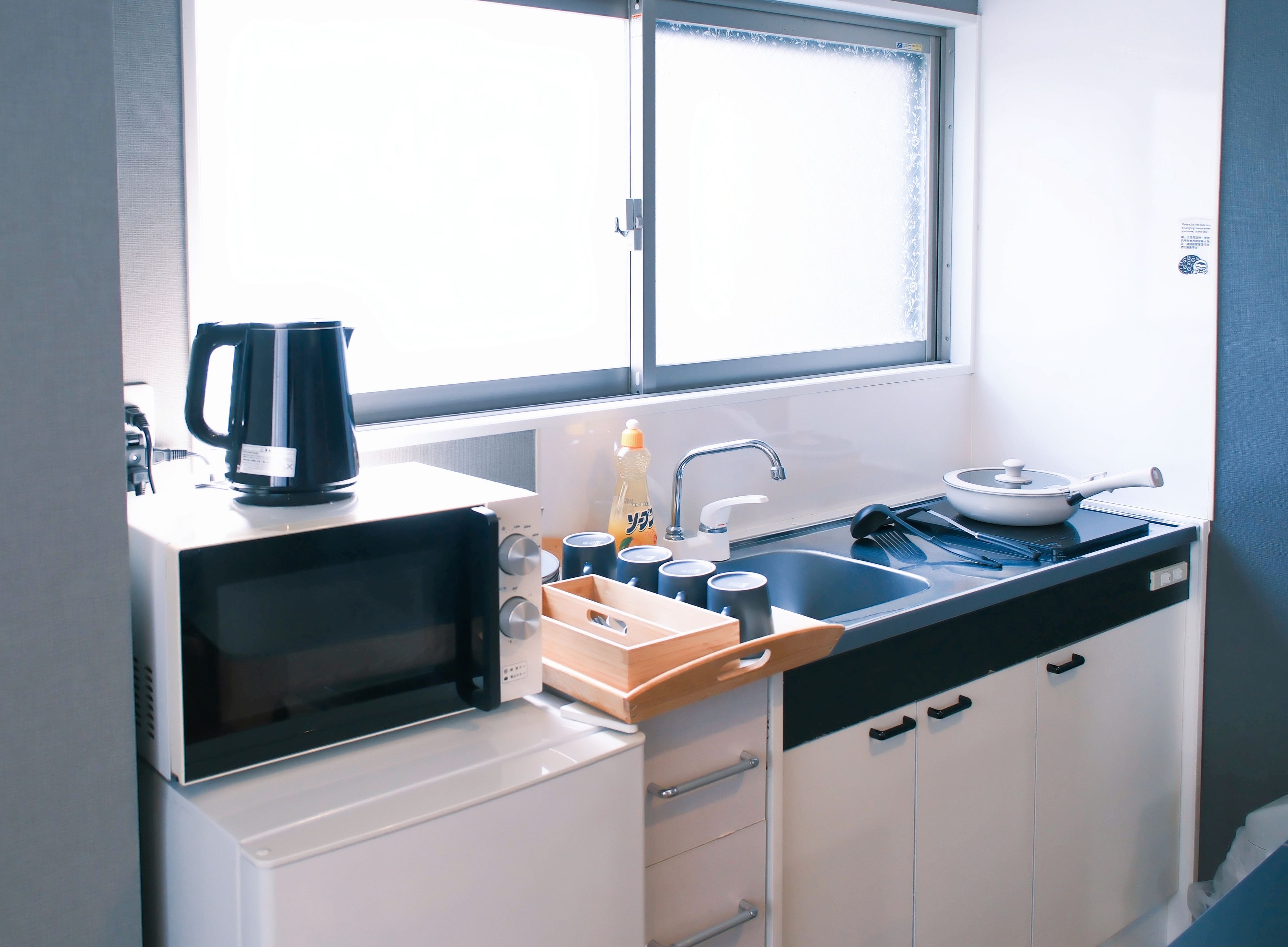 A compact kitchen with a microwave, kettle, sink, and a window above the counter.