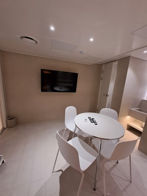 A modern room with a round table and four white chairs, a flat-screen TV mounted on the wall, and recessed ceiling lighting.