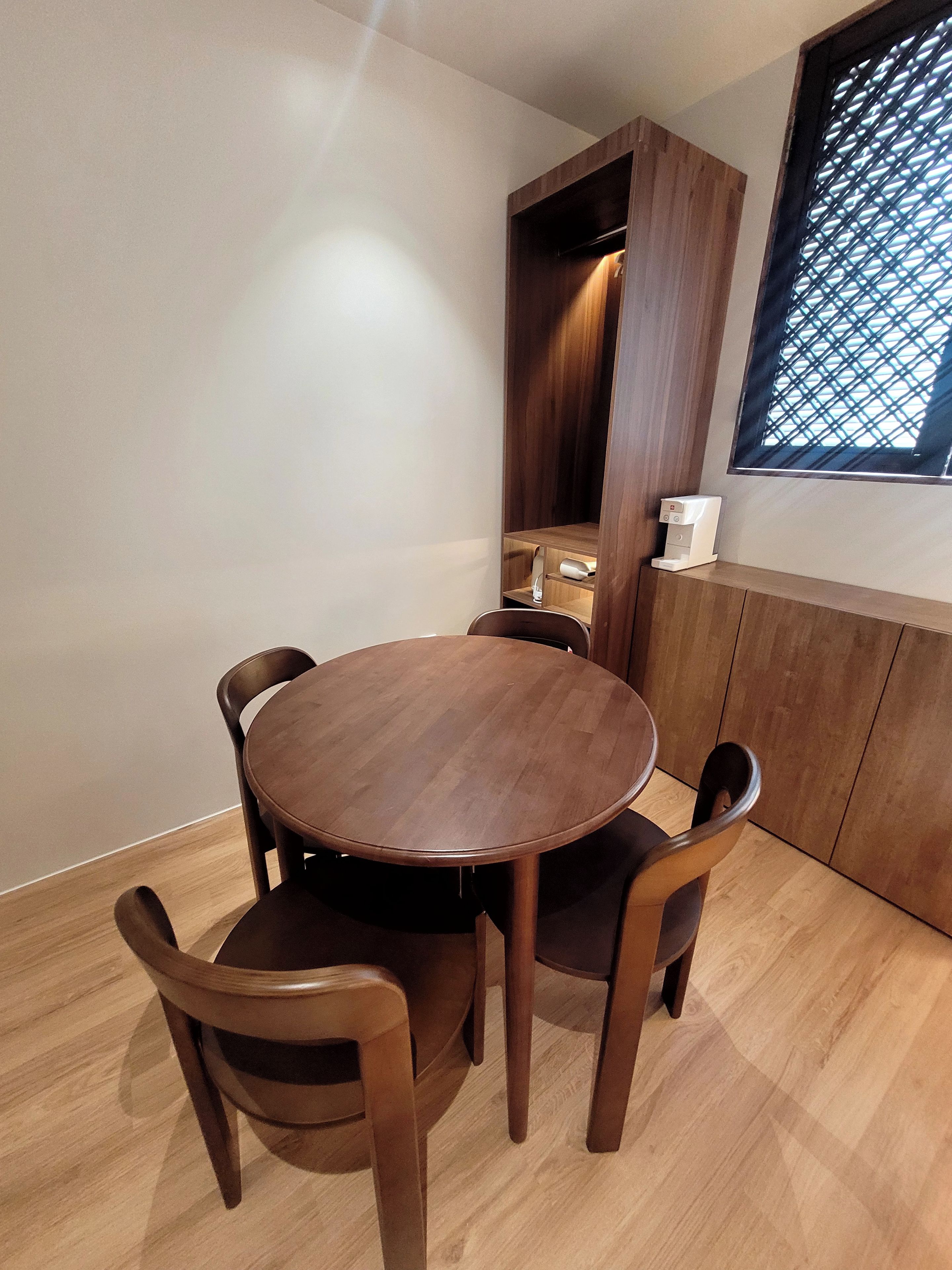A dining area with a round wooden table and four chairs, a built-in wooden cabinet, and a window with a lattice design.