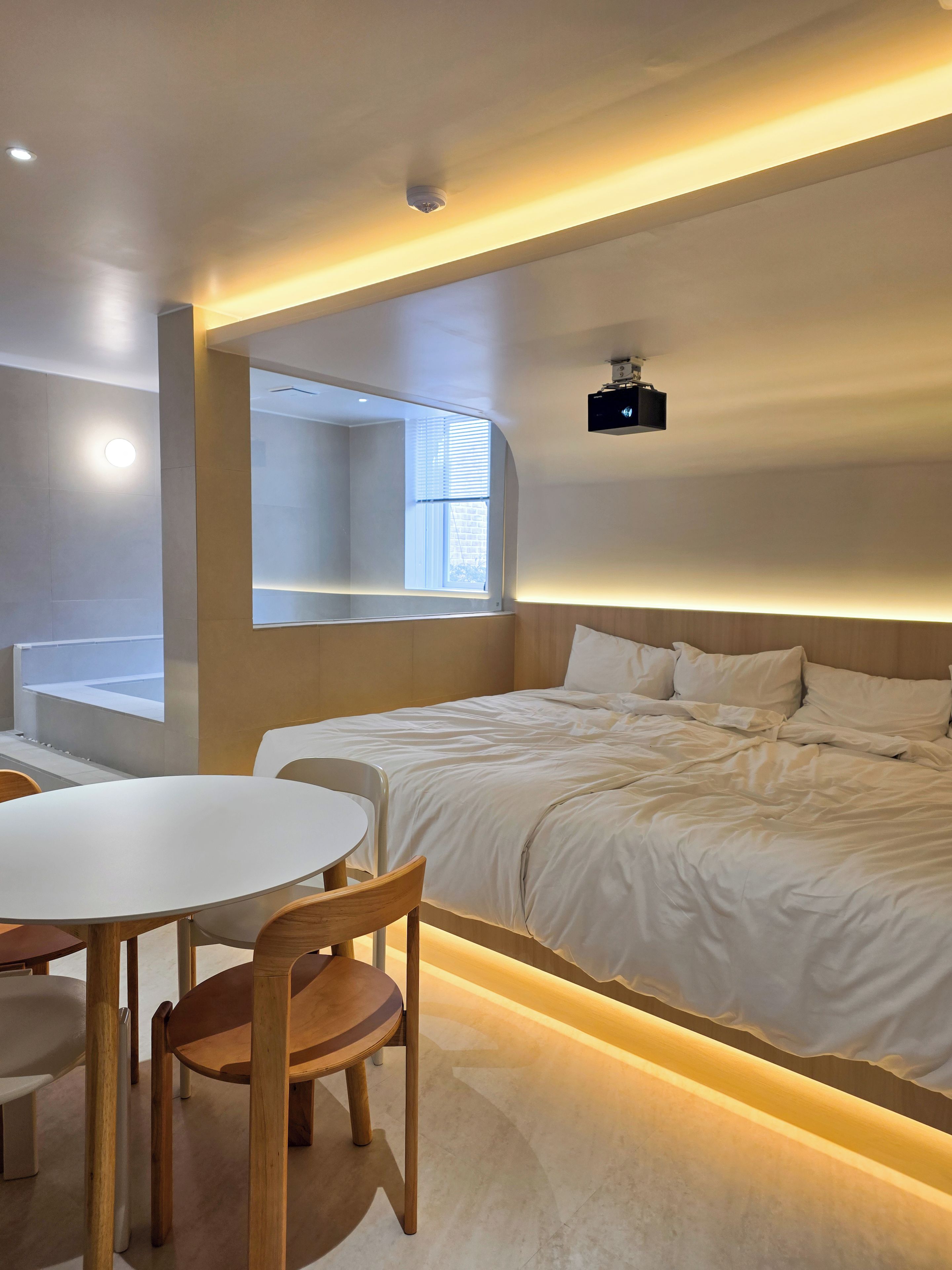 A modern hotel room with a neatly made bed, a round dining table with chairs, and a large window providing natural light.