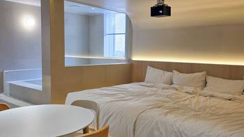 A modern hotel room with a neatly made bed, a round dining table with chairs, and a large window providing natural light.
