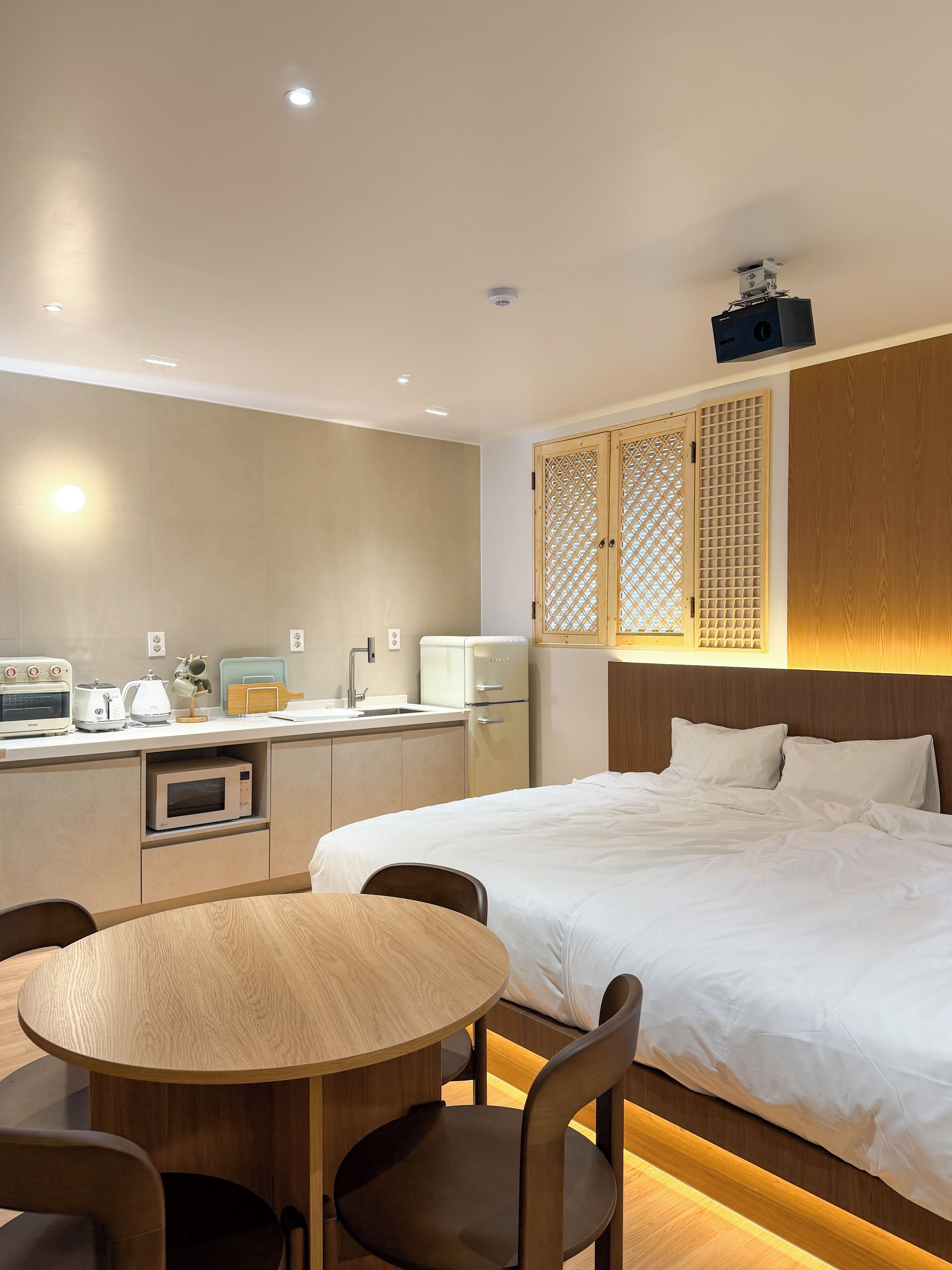 A hotel room with a bed, a dining table with chairs, a kitchenette, and a wooden headboard.