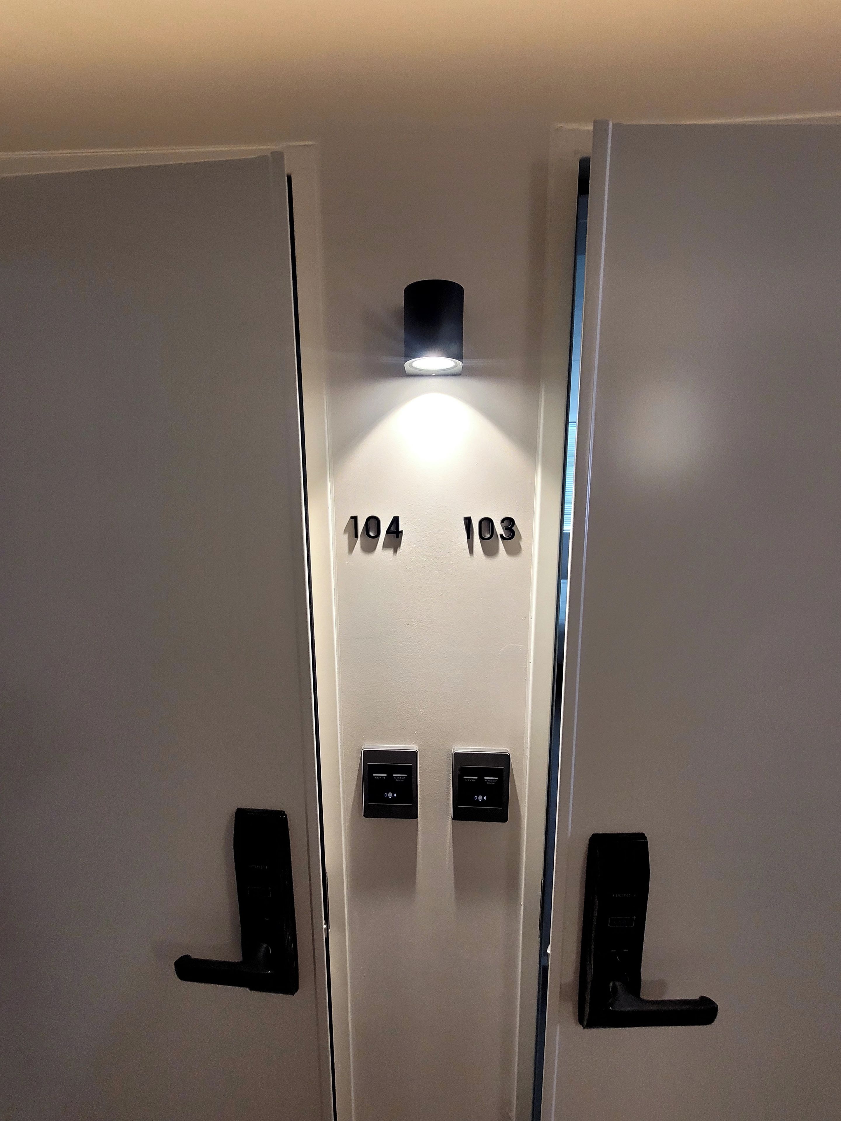 A hotel room with a door marked with numbers 104 and 103, indicating room numbers. There are two black electronic devices on the door, likely for room access or security.