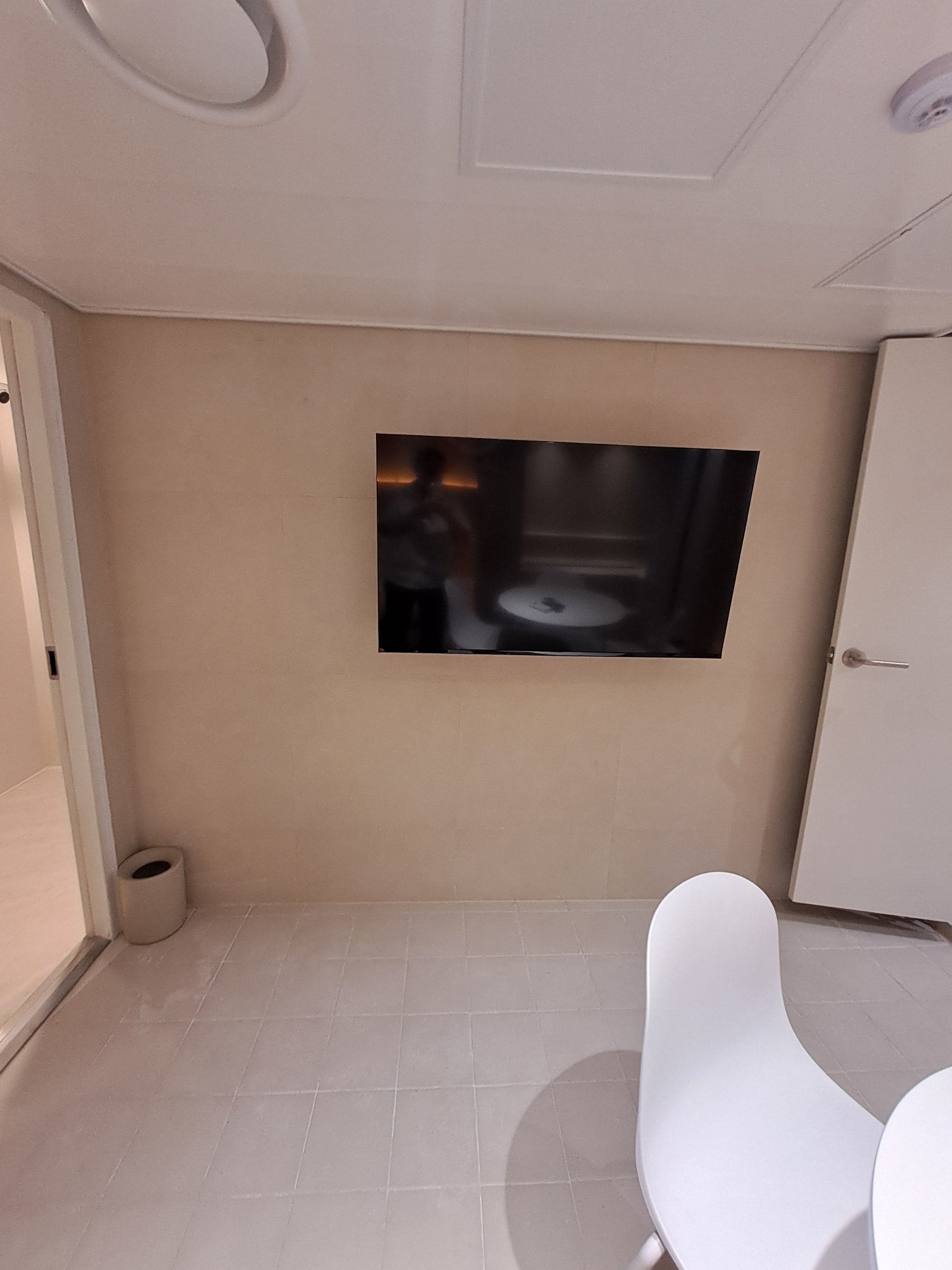 A modern room with a flat-screen TV mounted on the wall, a white chair, and a small trash bin.