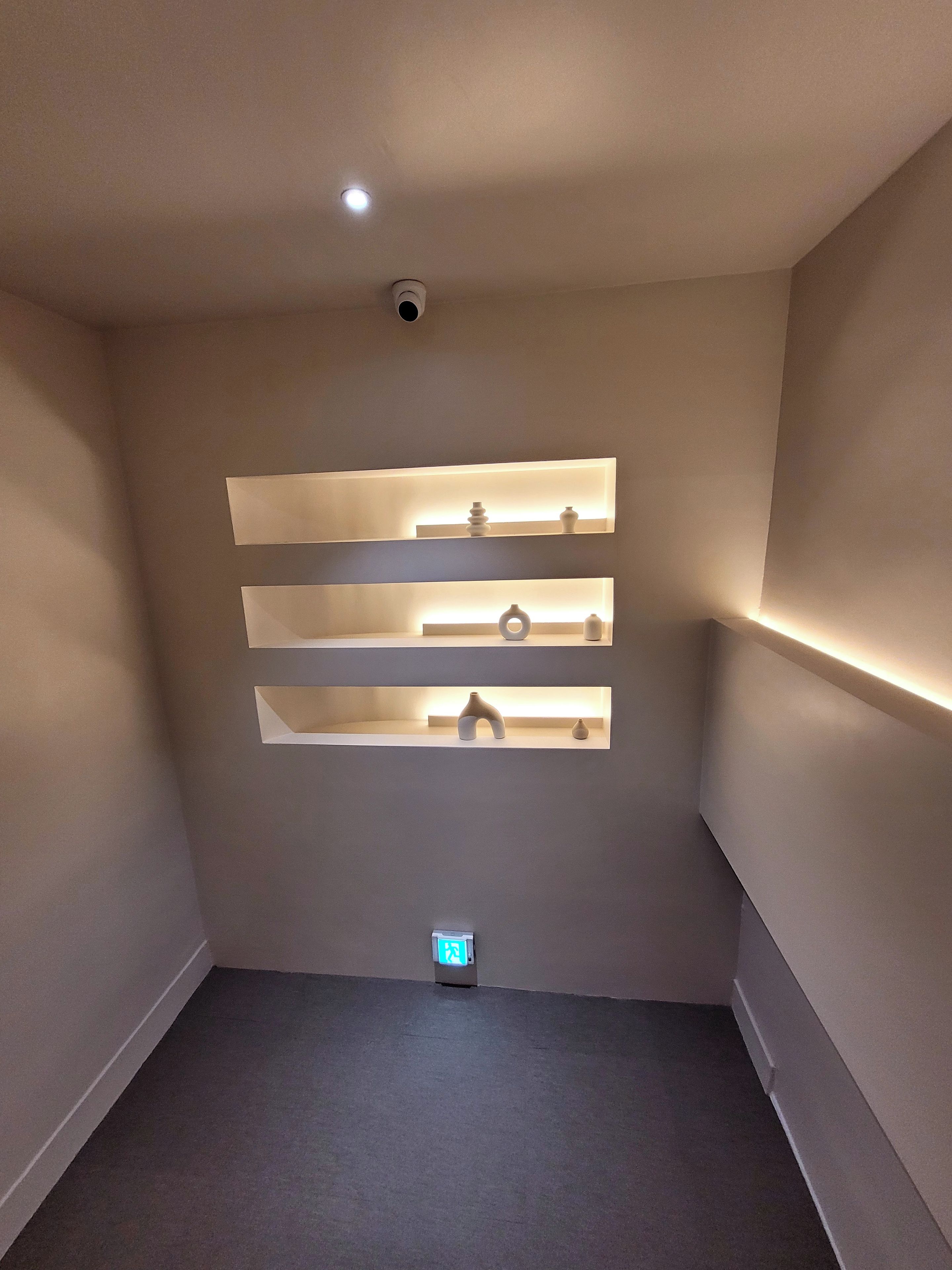 A room with a recessed shelf unit against the wall, a ceiling-mounted security camera, and a digital clock on the floor.