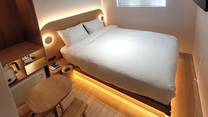 Un dormitorio moderno con una cama, mesita de noche, lámpara y un pequeño banco de madera.