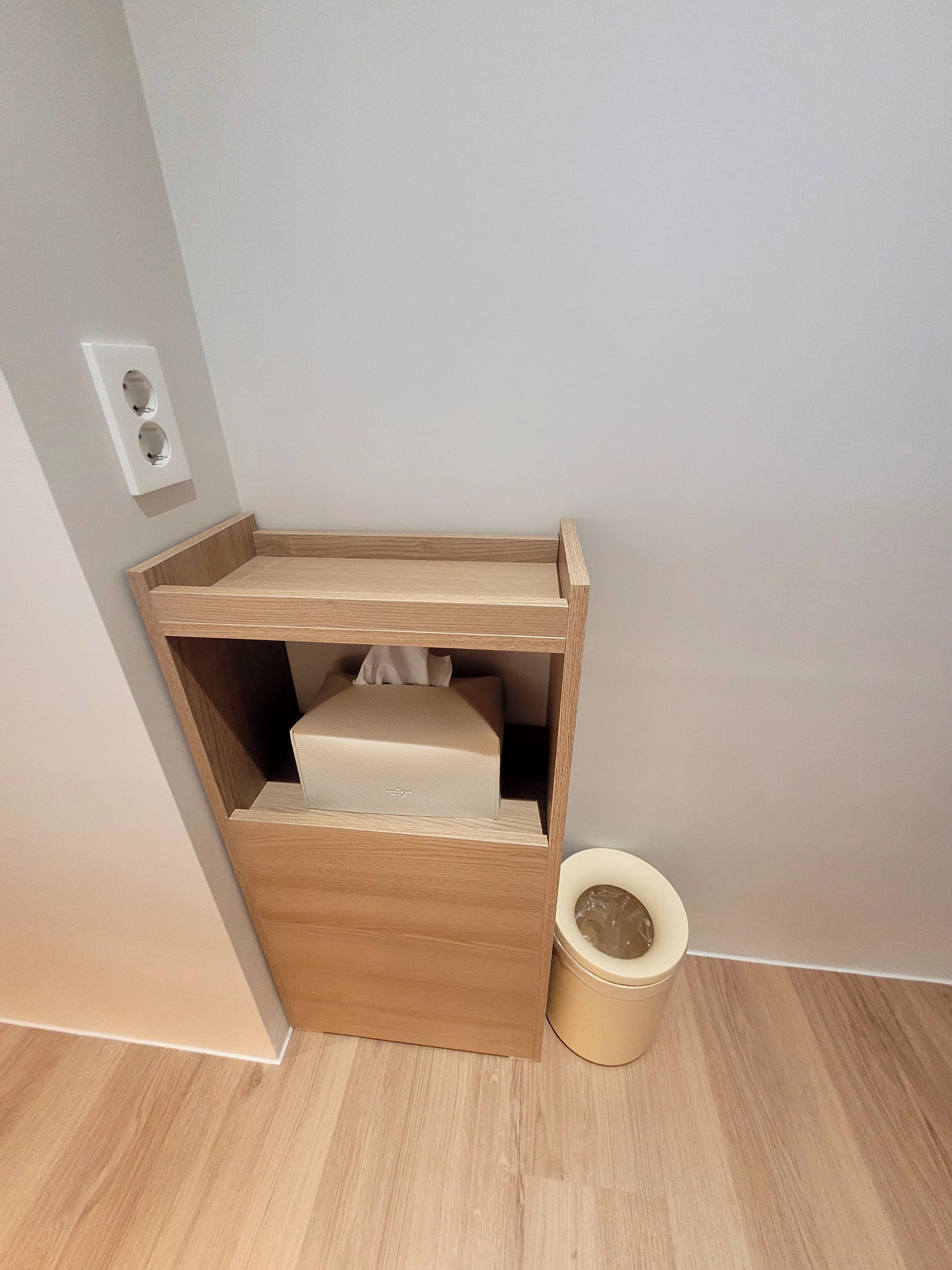 Una esquina de una habitación con un armario de madera, un cesto de basura pequeño y un enchufe de pared.