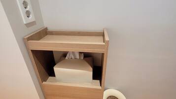 Una esquina de una habitación con un armario de madera, un cesto de basura pequeño y un enchufe de pared.
