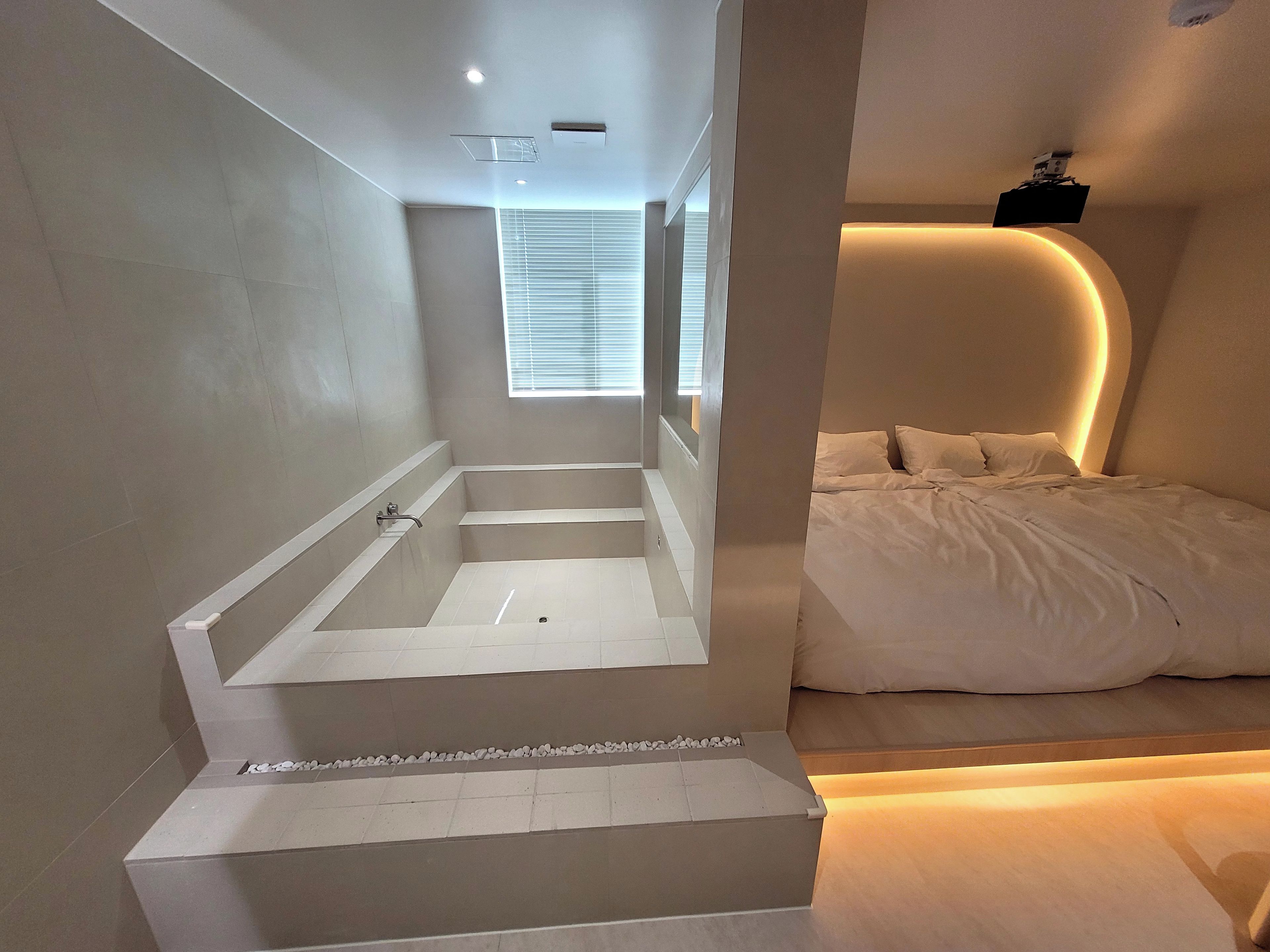 Une salle de bain moderne avec une baignoire, une douche et des toilettes. La pièce adjacente contient un lit avec du linge de lit blanc.