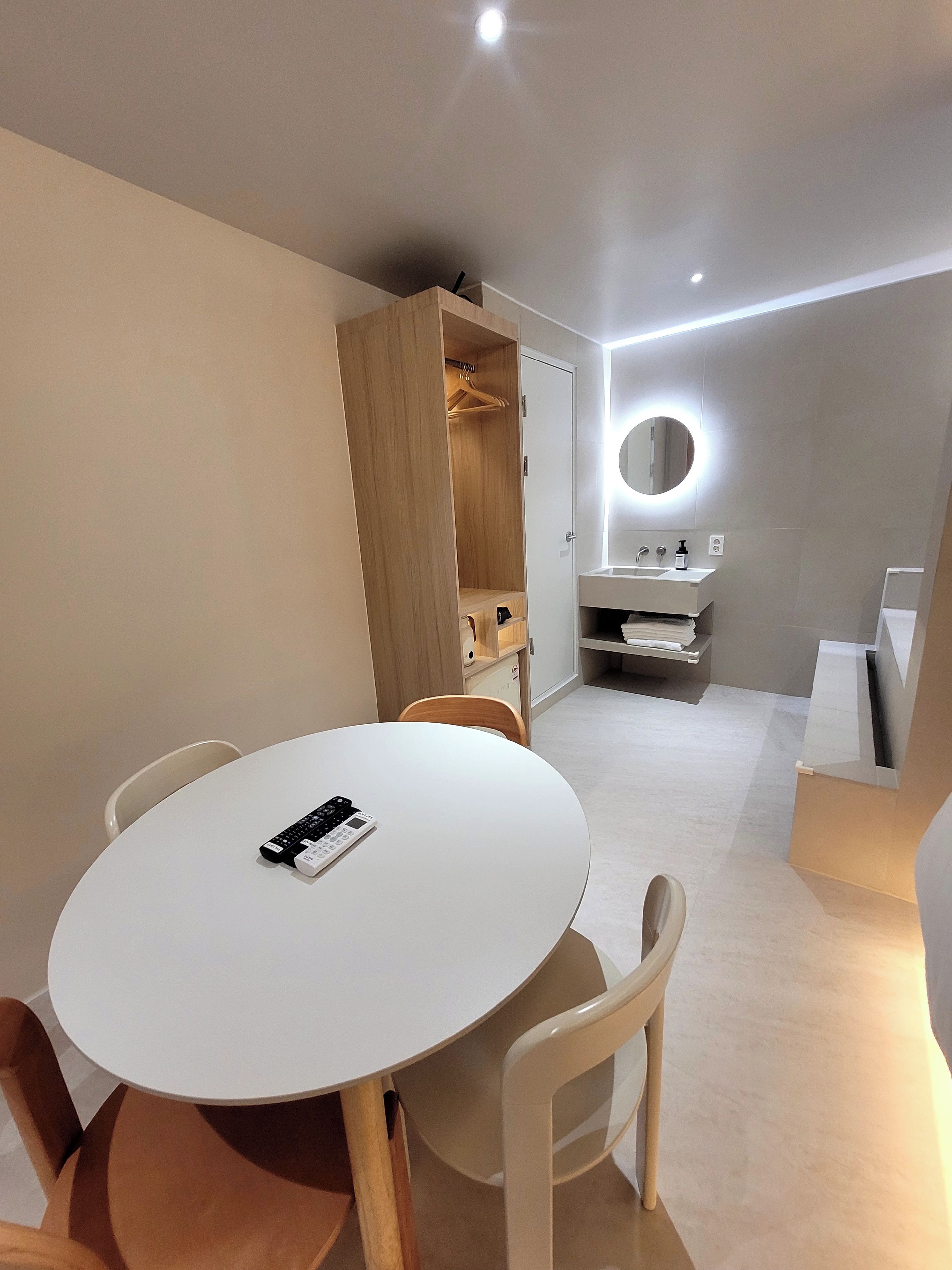 A modern, well-lit room with a round table, chairs, a wooden wardrobe, a mirror with lights, and a small sink.