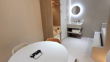 A modern, well-lit room with a round table, chairs, a wooden wardrobe, a mirror with lights, and a small sink.