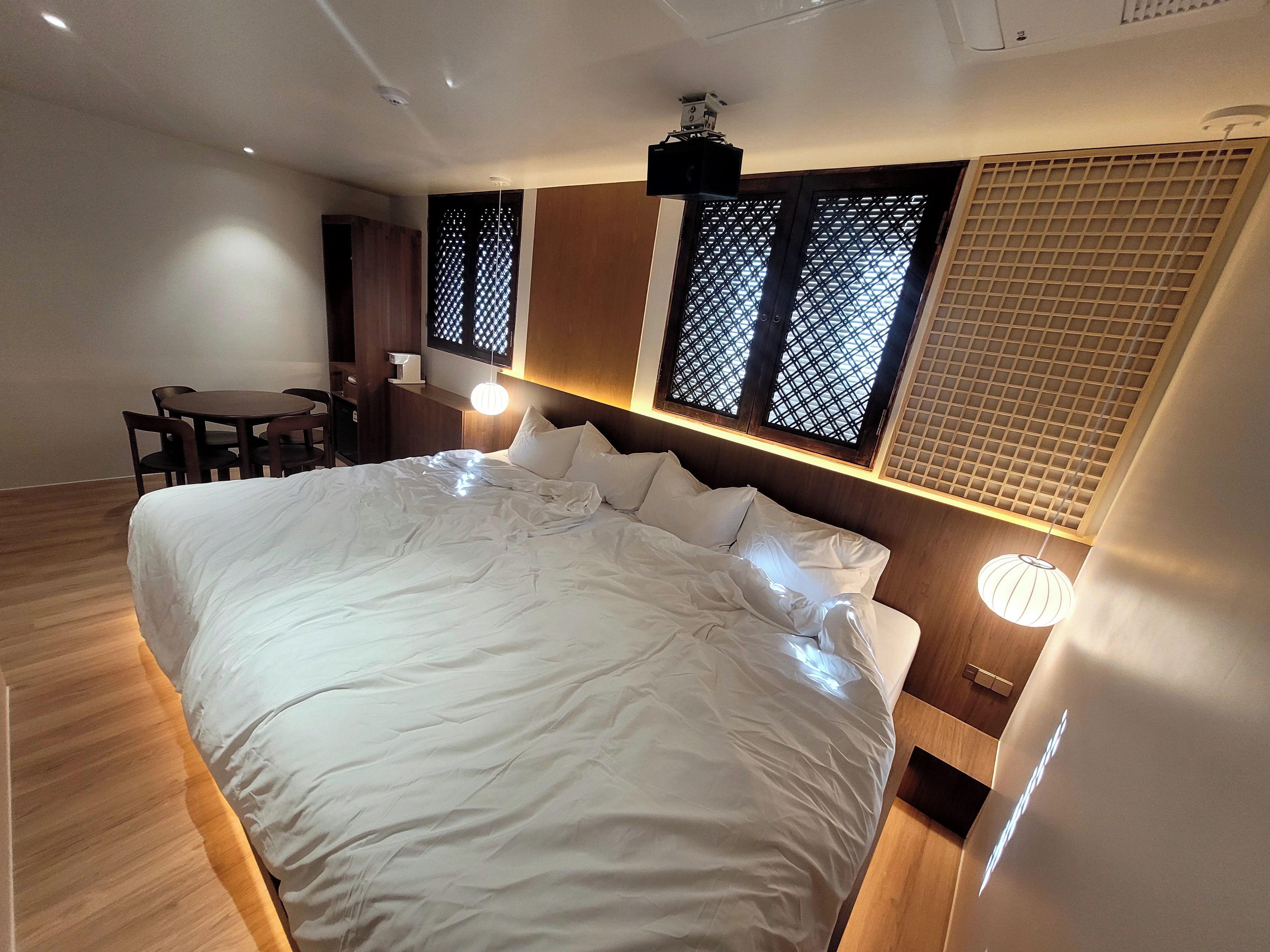 A hotel room with a large bed, a desk, and a chair. There are windows with wooden frames and a ceiling fan.